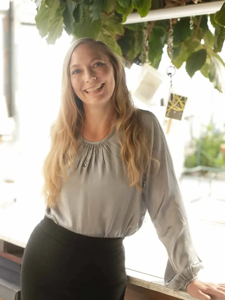 Linda Schraysshuen lächelt in die Kamera, mit langen, blonden Haaren, einer seidig glänzenden Bluse und einer entspannten Haltung vor einem hellen Fenster mit Pflanzen im Hintergrund.