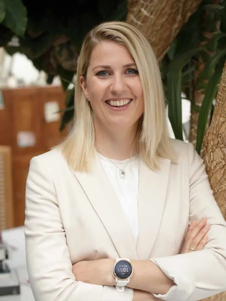 Anja Preuß steht mit verschränkten Armen vor einem Baum im Innenraum, trägt einen hellen Blazer und lächelt selbstbewusst in die Kamera.
