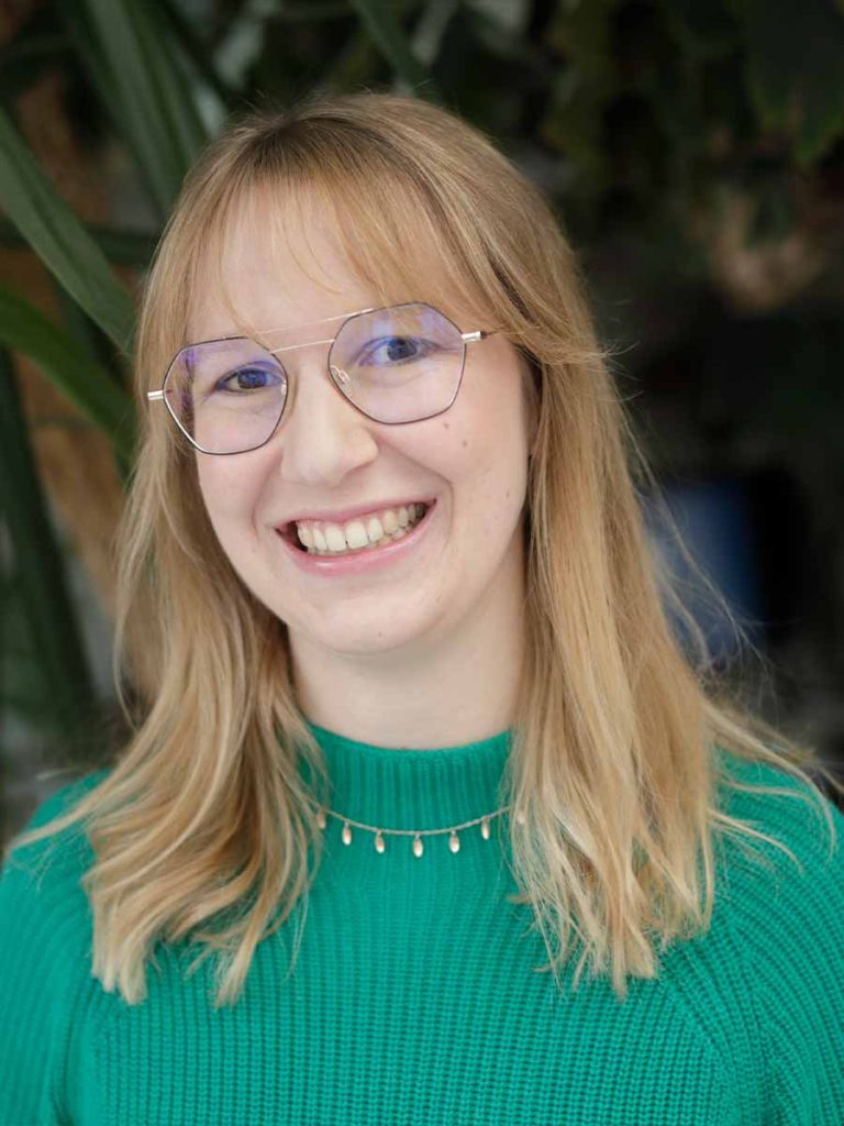 Anna-Lena Königbauer lächelt in die Kamera. Sie trägt eine runde Brille, einen grünen Strickpullover und eine zarte Halskette. Der Hintergrund ist natürlich und dezent gehalten.