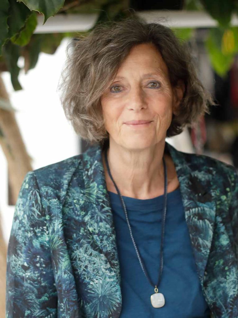 Brigitte Herkert blickt ruhig in die Kamera, mit halblangen, leicht gewellten Haaren und einem Blazer im grünen Pflanzenmuster. Sie trägt ein dunkelblaues Shirt und eine lange Kette mit Anhänger.