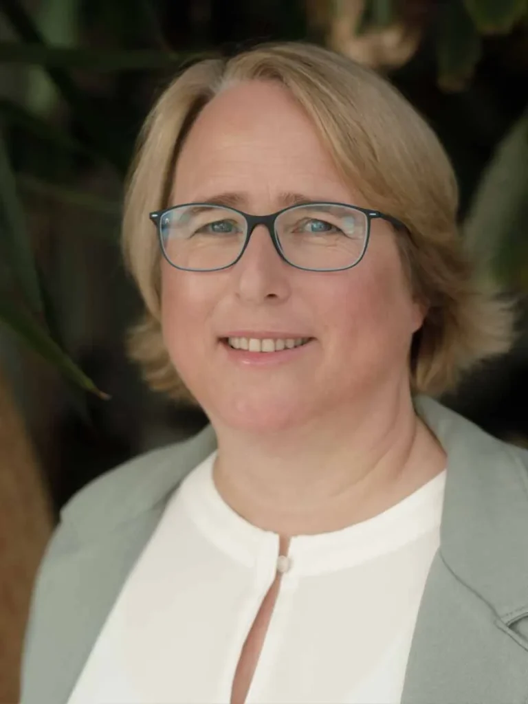 Doris Rudolf lächelt in die Kamera, trägt eine Brille mit dunklem Rahmen, eine helle Bluse mit kleinem Stehkragen und ein graugrünes Jackett. Der Hintergrund ist weich und natürlich gehalten.