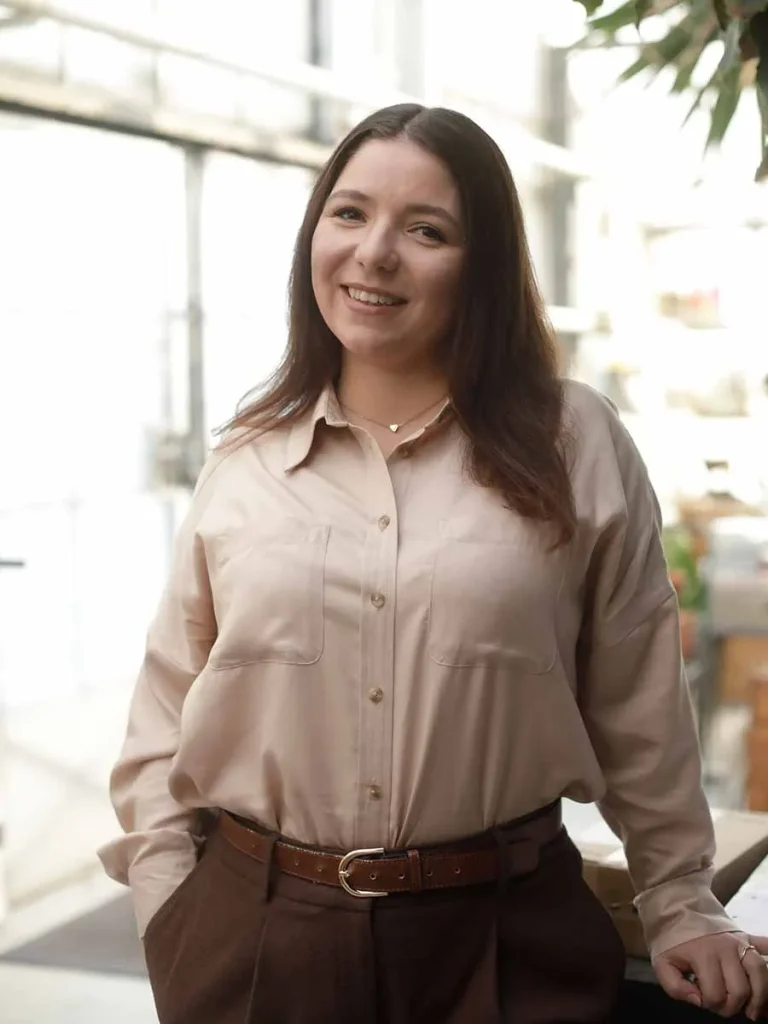 Magdalena Blumenfelder steht entspannt mit den Händen in den Hosentaschen und lächelt in die Kamera. Sie trägt eine beigefarbene Bluse und eine braune Hose mit Gürtel, im Hintergrund ist ein heller Arbeitsraum zu sehen.