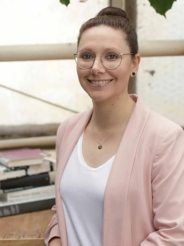 Meike Ganschiniet steht lächelnd vor einem Bücherstapel. Sie trägt einen rosafarbenen Blazer, ein weißes Shirt und eine runde Brille. Ihre Haare sind zu einem Dutt gebunden.