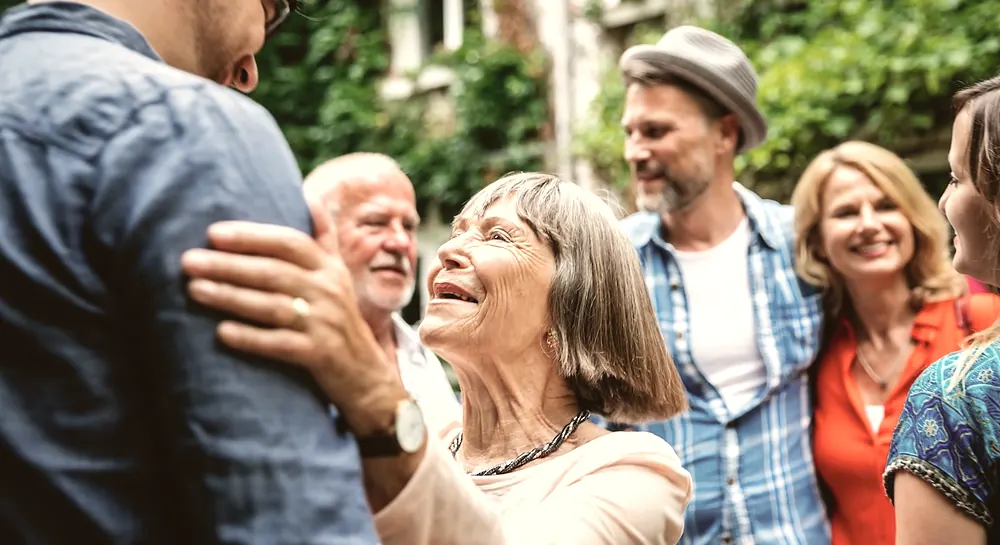 Eine ältere Frau begrüßt eine jüngere Person herzlich. Weitere Menschen verschiedener Generationen stehen lächelnd zusammen und tauschen sich im Freien aus